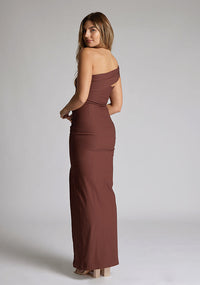 Back quarter image of a blonde 
model wearing a  Chocolate Maxi Dress with an asymmetric neckline and one-sleeve design and front skirt split adds allure.The design features Vesper Pearson Chocolate Bardot Maxi Dress