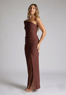Front quarter image of a blonde 
model wearing a  Chocolate Maxi Dress with an asymmetric neckline and one-sleeve design and front skirt split adds allure. The design features Vesper Pearson Chocolate Bardot Maxi Dress