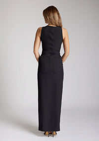 Back image of blonde model wearing a black asymmetrical detail maxi dress with a bold asymmetric neckline, front leg split and center back zip fastening. The design featured is the Vesper Polly Black Asymmetrical Detail Maxi Dress