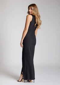 Back quarter image of blonde model wearing a black asymmetrical detail maxi dress with a bold asymmetric neckline, front leg split and center back zip fastening. The design featured is the Vesper Polly Black Asymmetrical Detail Maxi Dress
