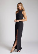 Front quarter image of blonde model wearing a black asymmetrical detail maxi dress with a bold asymmetric neckline, front leg split and center back zip fastening. The design featured is the Vesper Polly Black Asymmetrical Detail Maxi Dress