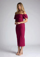 Front quarter image of a blonde model wearing a berry midaxi dress with a bardot neckline gracefully frame off shoulders, while the back zip ensures a seamless silhouette. The dress  features is the Vesper Quinton Berry Midaxi Dress