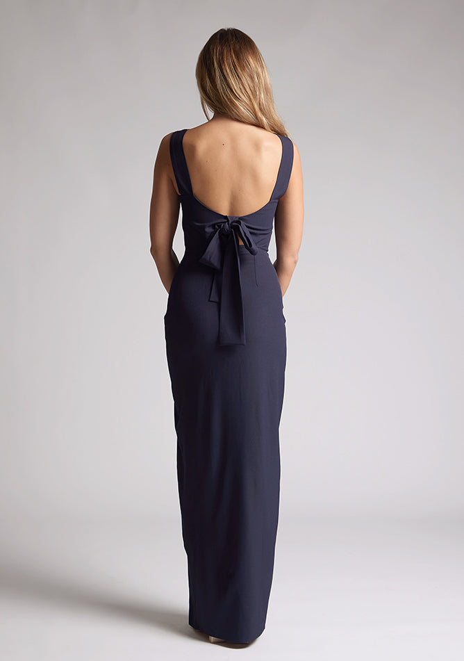 Back  image of a blonde model wearing a navy bow back maxi dress with a v neckline adds a touch of allure, front skirt split, while the bodycon fit flatters your figure elegantly and adding a touch of glamour. The design featured is the Vesper Reese Navy Bow Back Maxi Dress