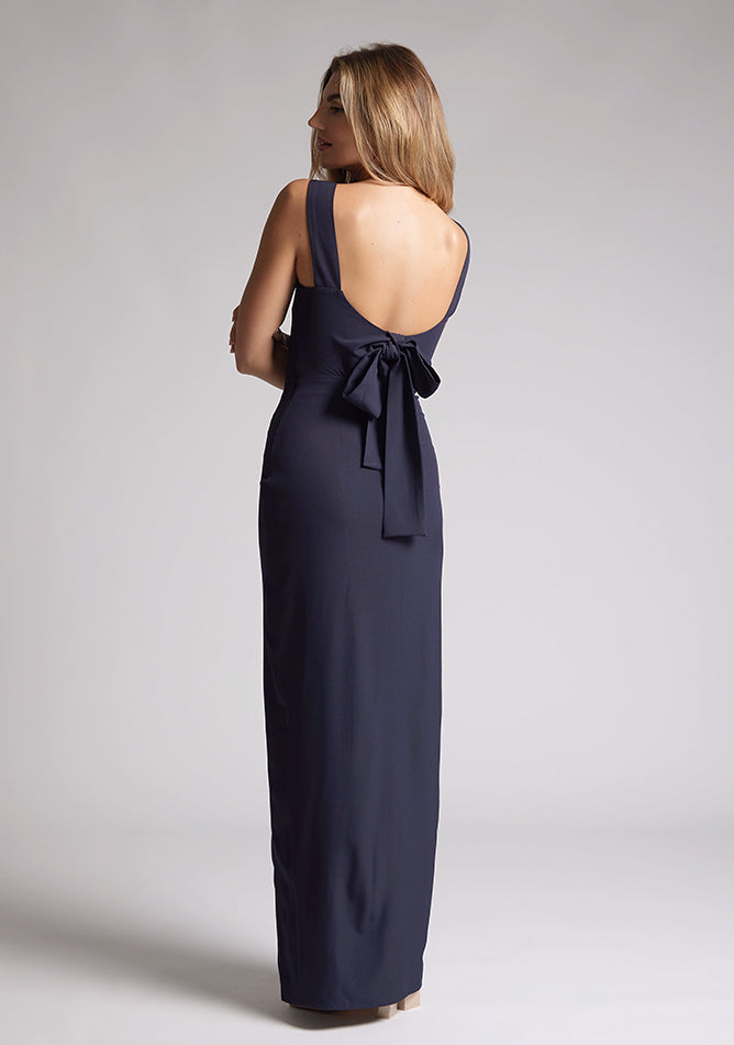 Back quarter image of a blonde model wearing a navy bow back maxi dress with a v neckline adds a touch of allure, front skirt split, while the bodycon fit flatters your figure elegantly and adding a touch of glamour. The design featured is the Vesper Reese Navy Bow Back Maxi Dress