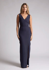 Front image of a blonde model wearing a navy bow back maxi dress with a v neckline adds a touch of allure, front skirt split, while the bodycon fit flatters your figure elegantly and adding a touch of glamour. The design featured is the Vesper Reese Navy Bow Back Maxi Dress
