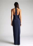 Back image of a blonde model wearing a navy maxidress with a v-plunging neckline, halter design and a front skirt split. The design featured is the Vesper Sedona Navy Maxi Dress