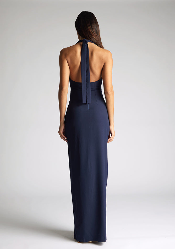 Back image of a blonde model wearing a navy maxidress with a v-plunging neckline, halter design and a front skirt split. The design featured is the Vesper Sedona Navy Maxi Dress