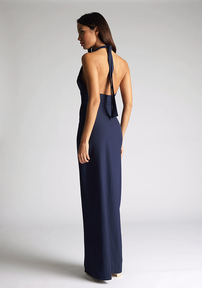 Back quarter  image of a blonde model wearing a navy maxidress with a v-plunging neckline, halter design and a front skirt split. The design featured is the Vesper Sedona Navy Maxi Dress