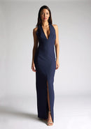 Front image of a blonde model wearing a navy maxidress with a v-plunging neckline, halter design and a front skirt split. The design featured is the Vesper Sedona Navy Maxi Dress