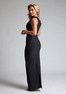Back quarter image of a blonde model wearing a black maxi dress with an sleeveless silhouette and add a modern twist. The design features is the Vesper Wyatt Black Asymmetric  Maxi Dress