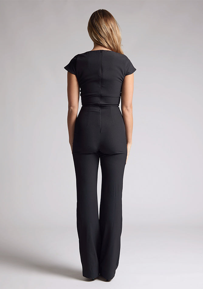 Back image of a model wearing a black jumpsuit with a square neckline and flattering v-wire design, this jumpsuit will have you feeling confident and stylish. The design features is the Vesper Zuri Black Wide Leg Jumpsuit